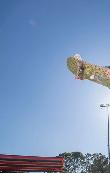 A skateboarder performs a high aerial grab above the concrete bowl at Motutawa Skatepark, New Zealand, against a clear blue sky. Spectators and a park setting are visible in the background. Photo credit: Convic.