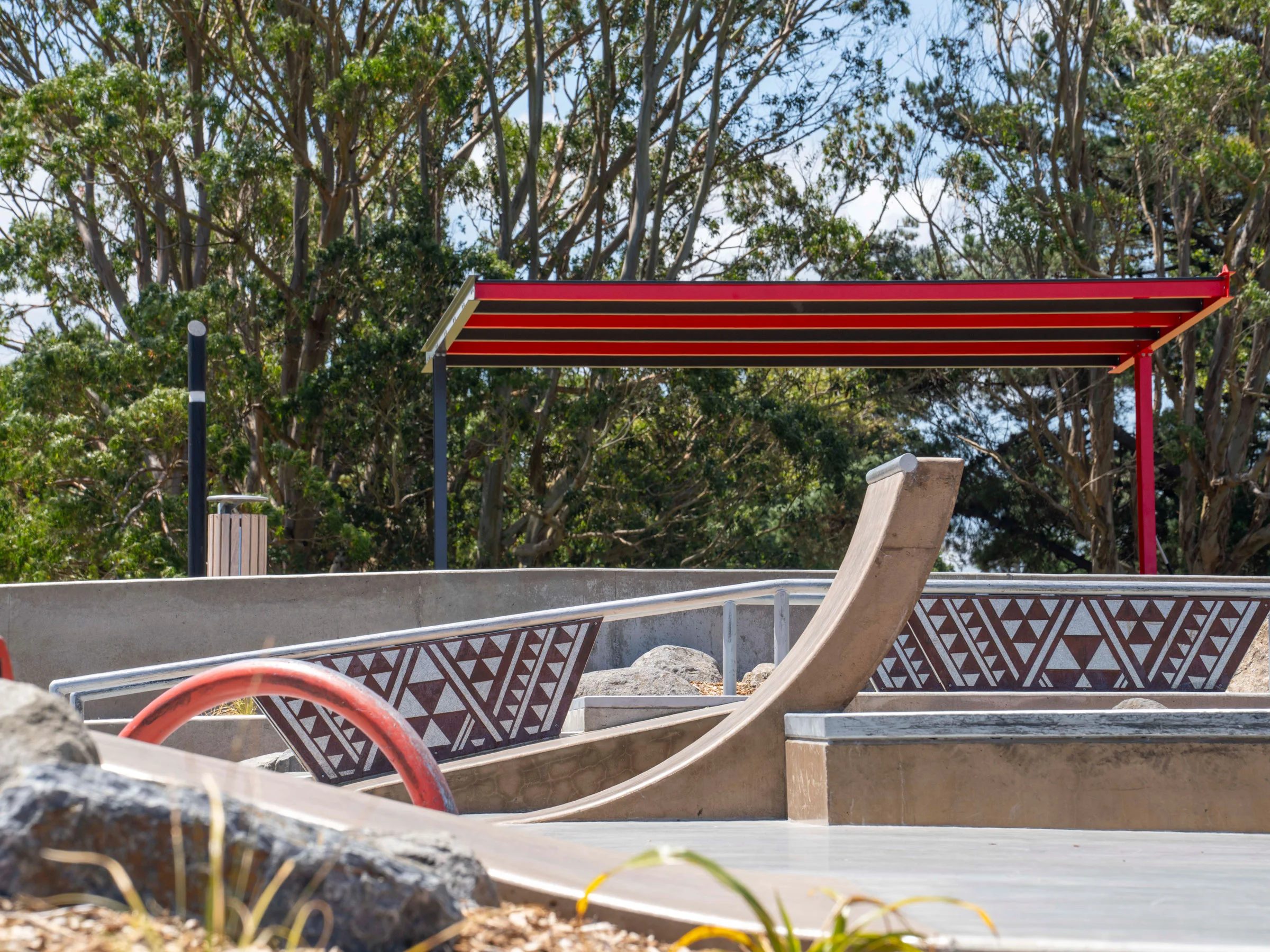 Close-up photograph of concrete skate ramp edges at Motutawa Skatepark, featuring traditional Māori geometric patterning inlaid into the concrete retaining walls. A red and striped steel shade canopy and native bush are visible in the background. Photo credit: Convic.