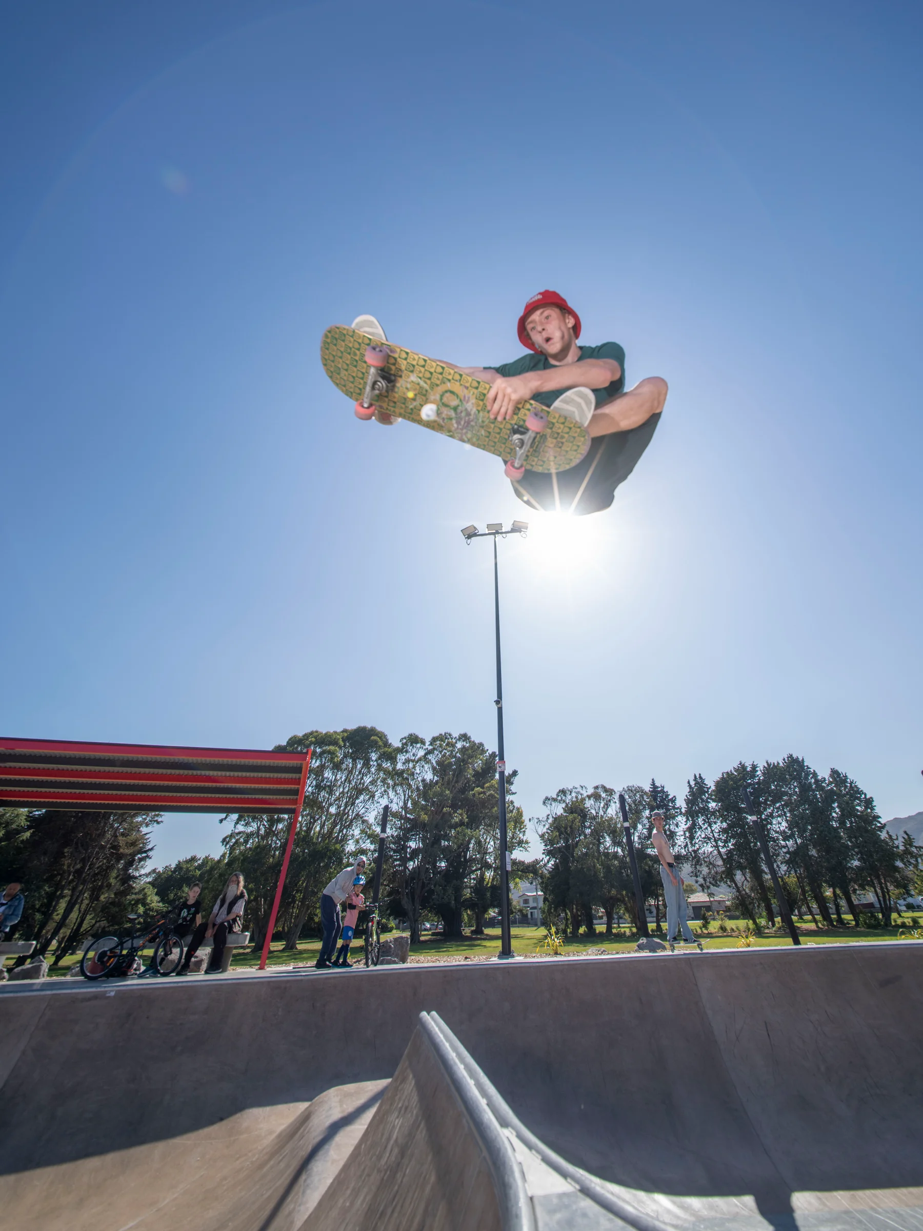 A skateboarder performs a high aerial grab above the concrete bowl at Motutawa Skatepark, New Zealand, against a clear blue sky. Spectators and a park setting are visible in the background. Photo credit: Convic.
